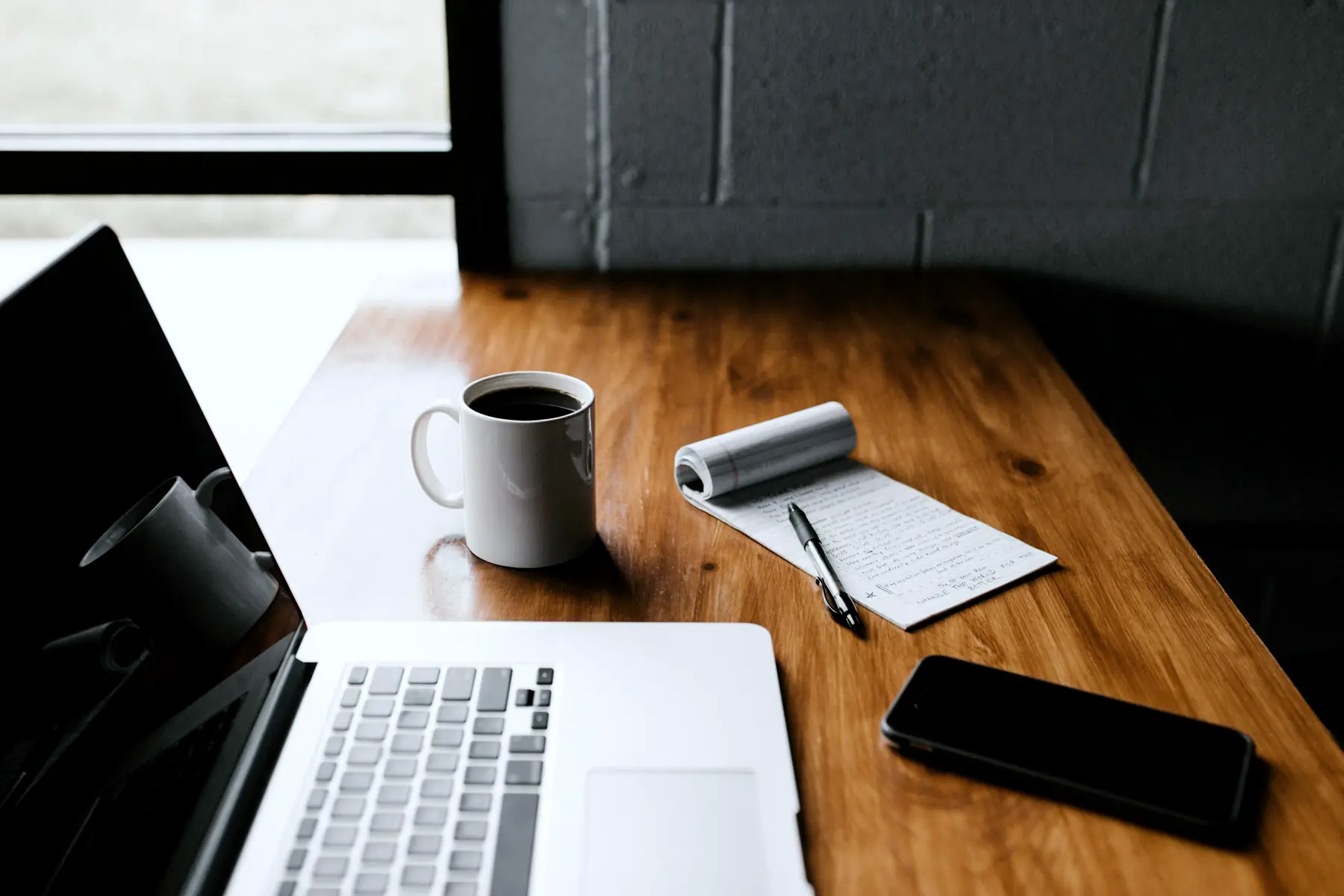 A laptop, a mug of coffee, a notepad and a phone on a wooden desk