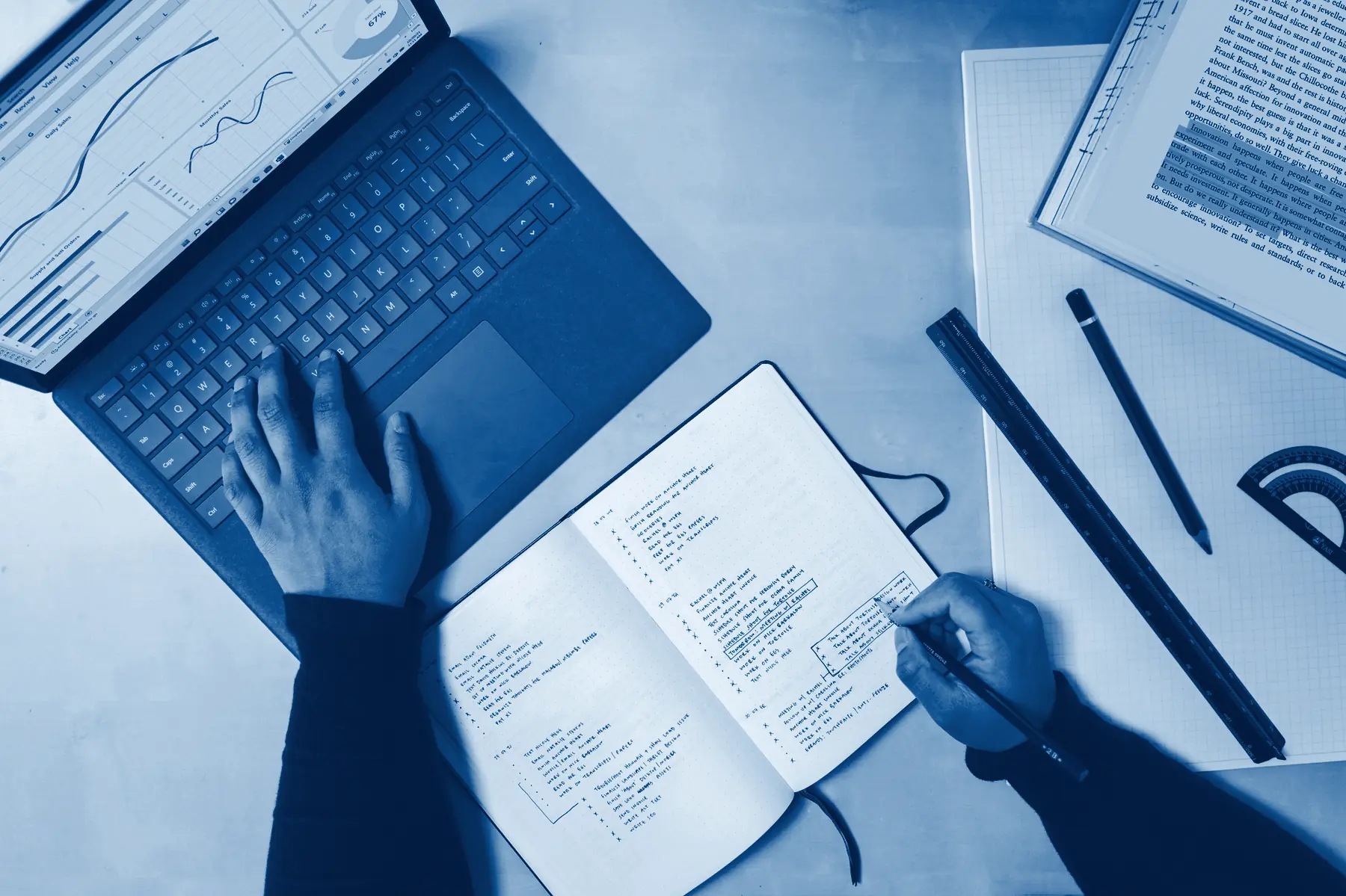 A hand on a laptop beside a notebook with a written plan, in Capdeva navy